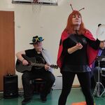 Czteroosobowy zespół muzyczny - od lewej instrument klawiszowy, gitara,  akordeon. Osoba prowadząca w stroju biedronki trzyma bębenek i pałeczkę w rękach.