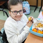 Dziewczynka na wózki siedzi przy stole. W rękach trzyma mandarynkę.  Przed nią leży pudełko z mandarynkami.