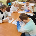 Grupa uczniów siedzą wokół stołu. Uczniowie pochylają się nad kartkami leżącymi na stole. trzymają w dłoniach długopisy.