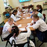 Grupa uczniów siedzą wokół stołu. Uczniowie pochylają się nad kartkami leżącymi na stole. Trzymają w dłoniach długopisy.
