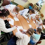 Grupa uczniów siedzi wokół stołu. Uczniowie pochylają się nad kartkami leżącymi na stole.