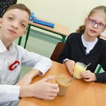 Uczeń i uczennica siedzą przy stole. trzymają w rękach kubki z budyniem.