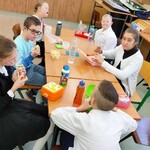 Uczniowie siedzą wokół stołu. Przed nimi stoją pudełka śniadaniowi i butelki z piciem.