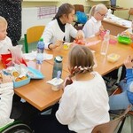 Uczniowie siedzą wokół stołu. Przed nimi stoją pudełka śniadaniowi i butelki z piciem.