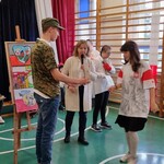Nauczycielka trzyma mikrofon. Obok stoi bokiem uczeń trzymający w ręku papierową torebkę oraz uczennica w koszulce z orłem.