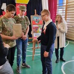 Trójka uczniów stoi naprzeciwko siebie, Jeden uczeń trzyma papierową torebkę. Z tyłu stoi nauczycielka z mikrofonem w dłoni.