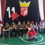Uczennica w czerwonej sukience na wózku trzyma mikrofon w dłoniach. Za nią na ławeczce siedzi pięciu uczniów w koszulkach i czapkach moro oraz czterech w strojach ułana.