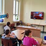 Trójka uczniów siedzi przy ławkach, zwróceni są w stronę ściany na której wisi telewizor i oglądają prezentację multimedialną o jesieni.