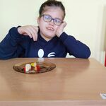Uczennica siedzi przy ławce. Prze nią stoi talerz, a na nim kawałki owoców. Dziewczynka po wyglądzie odgaduje co to za owoc.