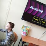 Uczeń siedzi na krześle przed laptopem i rozwiązuje na komputerze na czas przykłady matematyczne. Na ścianie za uczniem wisi telewizor na którym wyświetlane są przykłady.