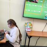 Uczennica siedzi na krześle przed komputerem rozwiązuje przykład matematyczny. Na ścianie znajduje się telewizor na którym widać rozwiązane przykłady.