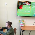 Uczeńsiedzi na krześle przed komputerem rozwiązuje przykład matematyczny. Na ścianie znajduje się telewizor na którym widać rozwiązane przykłady.