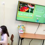 Uczennica siedzi na krześle przed komputerem rozwiązuje przykład matematyczny. Na ścianie znajduje się telewizor na którym widać rozwiązane przykłady.