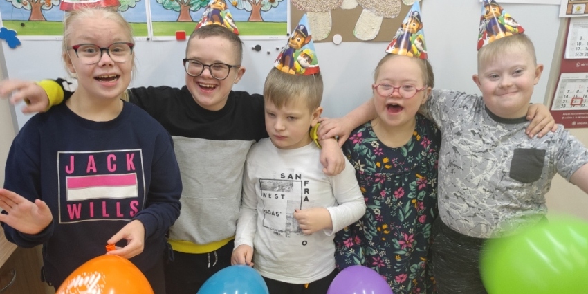 Grupa uśmiechniętych uczniów stoi przed tablicą,W ręku trzymają kolorowe balony.