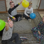 Uczniowie w urodzinowych czapeczkach siedzi na podłodze, w ręku trzymają balony.