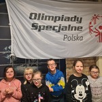Szóstka dzieci stoi na tle ściany z cegły i pozuje do zdjęcia. Nad nimi wisi biały baner z symbolem Olimpiady Specjalne Polska.