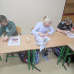 Uczniowie siedzą w ławkach. Pochylają się nad kartami pracy leżącymi przed mini na stole.