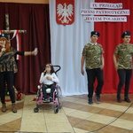 Dwóch uczniów oraz nauczycielka w koszulkach moro i beretach na tle dekoracji - biało-czerwona flaga, godło i napis. W środku dziewczynka na wózku.