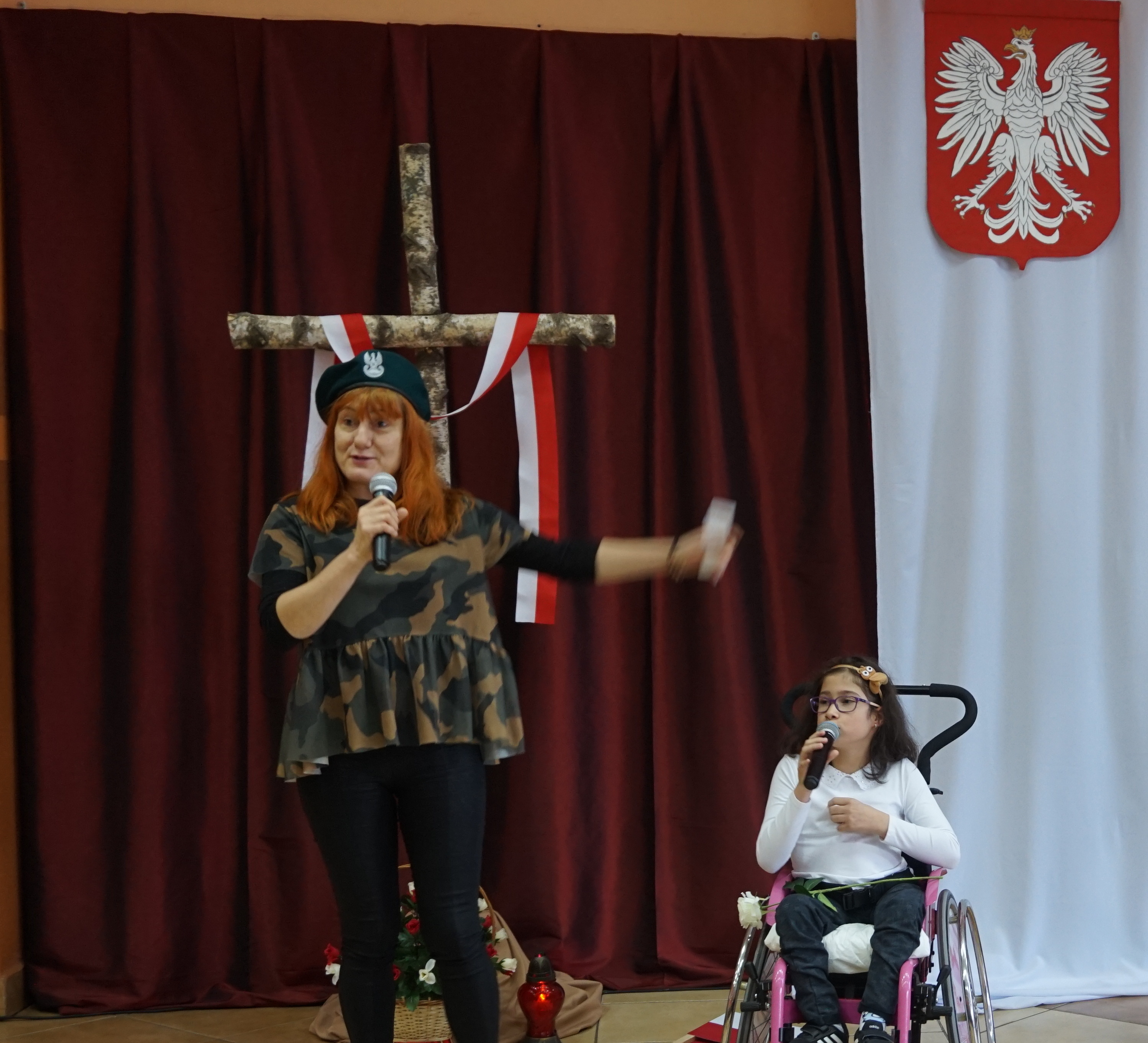 Dwóch uczniów oraz nauczycielka w koszulkach moro i beretach na tle dekoracji - biało-czerwona flaga, godło i napis. W środku dziewczynka na wózku.