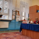 Uczeń czyta z kartki trzymanej w rękach. Jury konkursu siedzi za stołem przykrytym niebieskim obrusem.