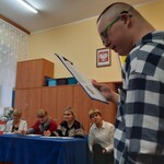 Uczeń czyta z kartki trzymanej w rękach. W tle siedzi za stołem czteroosobowe jury konkursu.
