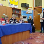 Nauczycielka stoi obok uczennicy czytającej z kartki. Po lewej jury konkursu siedzi za stołem przykrytym niebieskim