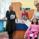 Na zdjęciu znajduje się dziewczynka na wózku, za nią stoi Pani Dyrektor, z lewej strony stoi Pani, która w ręku trzyma nagrodę, a z tyłu znajduje się jury.