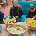 Grupa uczniów siedzi przed stołem, na którym stoją miski z przekąskami, picie i szklanki.