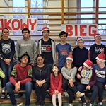 Grupa uczniów w mikołajowych czapkach z nauczycielką na tle drabinek i napisu Mikołajkowy bieg.
