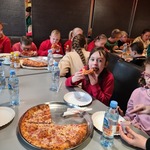 Uczniowie siedzą przy stołach w pizzerii. Przed nimi stoją butelki z wodą i talerze pizzą.