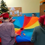 Przebrani uczniowie z nauczycielami bawią się kolorową chustą animacyjną. Na dalszym planie choinka.