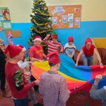 Przebrani uczniowie z nauczycielami bawią się kolorową chustą animacyjną. Na dalszym planie choinka.