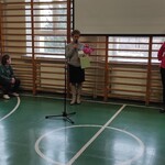 Na środku zdjęcia stoi nauczycielka z kwiatami, obok pani dyrektor. po bokach siedzą na ławeczkach uczniowie i nauczyciela.