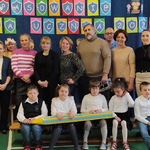 Uczniowie siedzą na ławeczce. Za nimi stoją rodzice i nauczycielki.