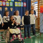 Uczennica na wózku śpiewa do mikrofonu. Za nią stoją uczniowie w odświętnych strojach.