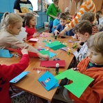 Grupa dzieci siedzi dookoła stołu na którym leżą materiały papiernicze. Nauczycielki obserwuja prace dzieci.