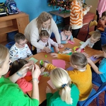 Dzieci siedzą wokół stołu na którym leżą materiały i przybory plastyczne. Nauczycielki obserwują prace dzieci. Jedna pochyla się nad dzieckiem.