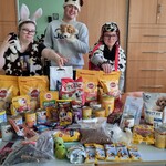 Trójka uczniów w przebraniu zwierząt stoi za stołem. Na stole leżą artykuły i smakołyki dla psów oraz sucha karma w workach i konserwy. 