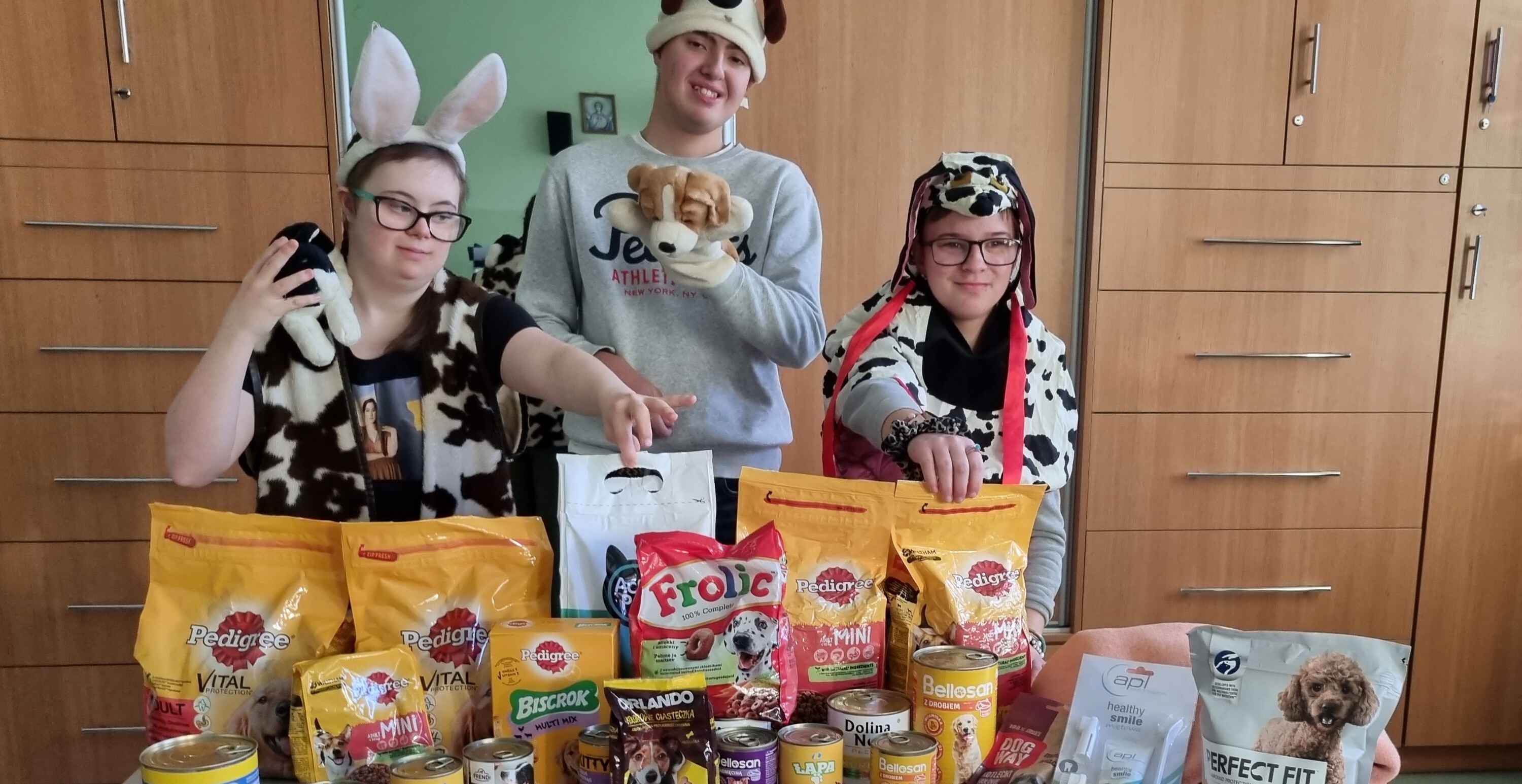 Trójka uczniów w przebraniu zwierząt stoi za stołem. Na stole leżą artykuły i smakołyki dla psów oraz sucha karma w workach i konserwy. 