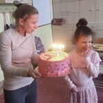 Nauczycielka trzyma przed uczennicą tort na którym są zapalone świeczki.