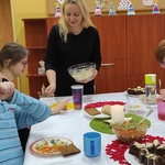 Uczniowie siedzą wokół stołu. Wychowawczyni podaje miskę z potrawą uczennicy.