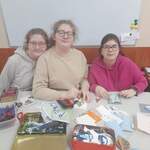 Przy stole pełnym pudełek z materiałami do świątecznych pocztówek siedzą trzy uczennice. Uśmiechają się i przytulają do siebie, przytrzymując leżące na stole kartki Bożonarodzeniowe.