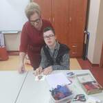 Uczeń siedzi przy stole pełnym pudełek z kreatywnymi materiałami plastycznymi. Przykleja elementy do świątecznej kartki. Za nim stoi nauczycielka i patrzy na jego pracę.