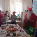 Wigilijny stół. Za nim stoją cztery osoby. Obok stołu stoi Mikołaj.