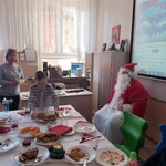 Wigilijny stół. Obok siedzi Mikołaj. Za stołem stoi nauczycielka i chłopiec nachylający się nad talerzem z potrawą.