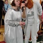 Nauczycielka i uczennica w białej tunice i wianku z piór śpiewają do mikrofonu.