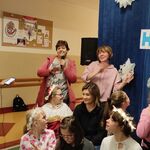 Uczniowie siedzą na ławeczkach. Z tyłu nauczyciele śpiewają kolędę.