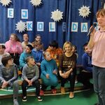 Uczniowie siedzą na ławeczkach na tle napisu Hej Kolęda. Nauczycielka mówi przez mikrofon.