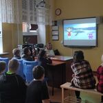 Uczniowie siedzą w ławkach i oglądają film wyświetlany na tablicy interaktywnej.