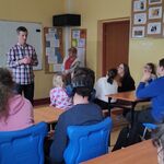 Uczniowie siedzą w ławkach i słuchają stojącego przed nimi nauczyciela.